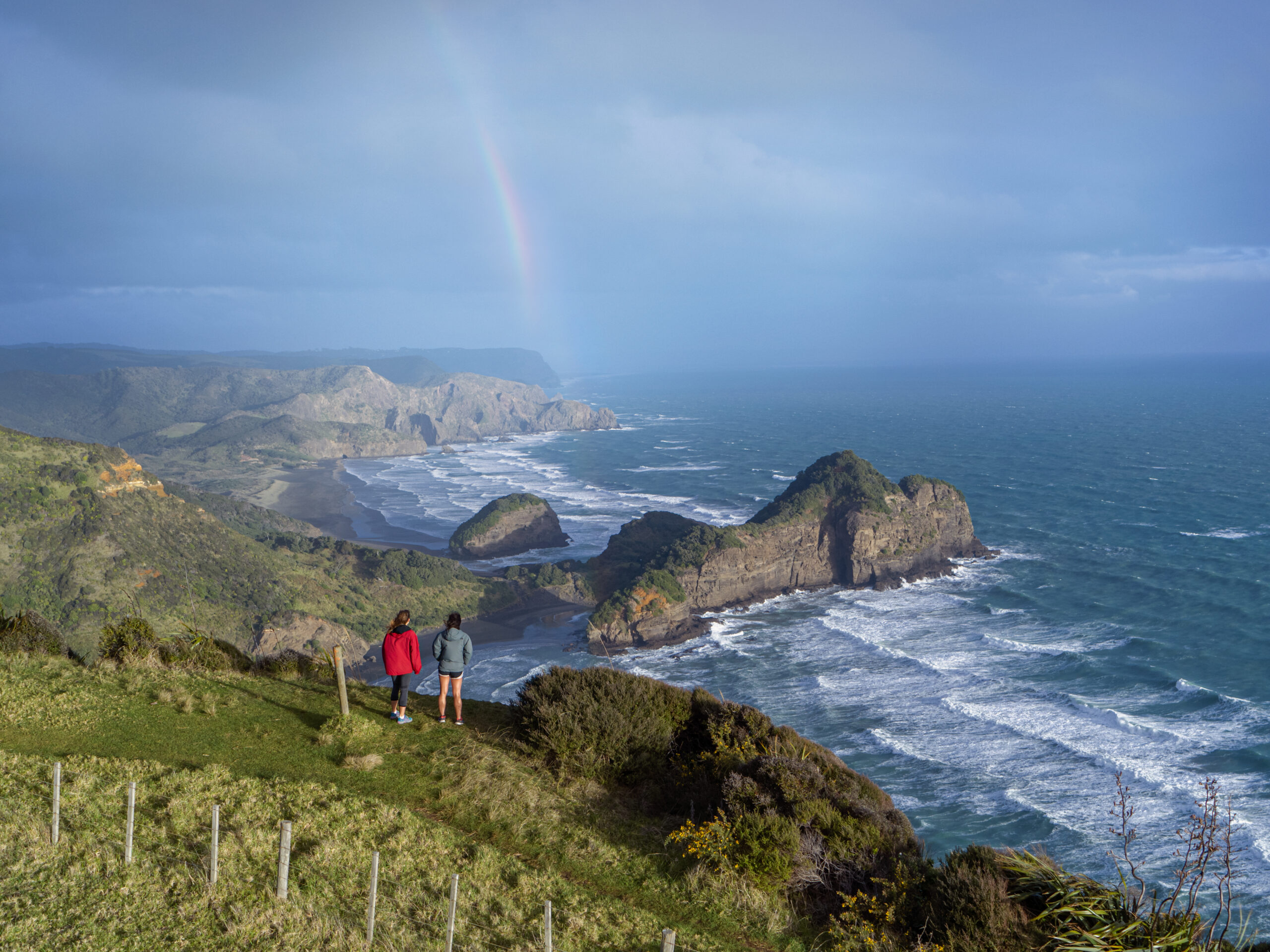 Two hikers on a clifftop looking out to the ocean with a rainbow at Aucklands West Coast